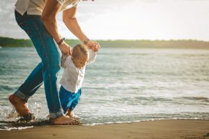 A child experiencing ocean waves for the first time, holding his father's hands and smiling